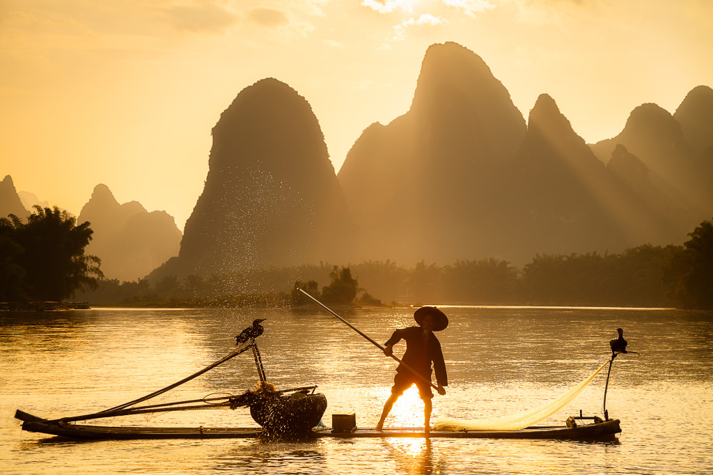 A Chinese fisherman on the Li River splashes water with an oar while Cormorants sit on the boat. Getting a sunset like this doesn't happen every day in this area.